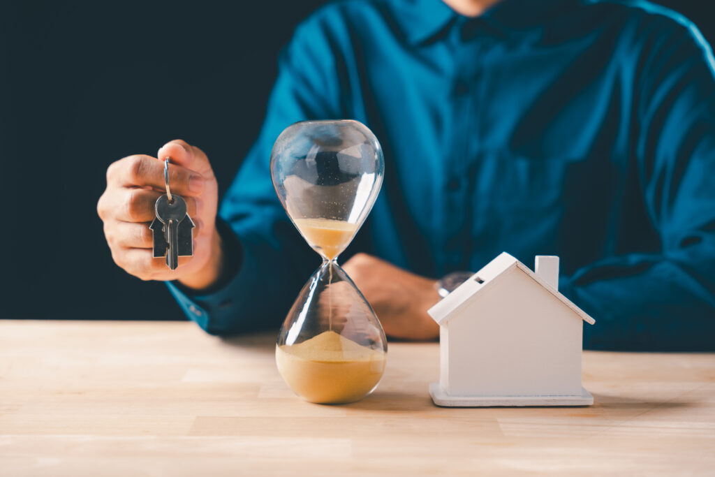 Hand holds real estate house keys, hourglass with falling sand marks time, symbolizing mortgage deadline, home buying, property loan concept for urgent housing investment.